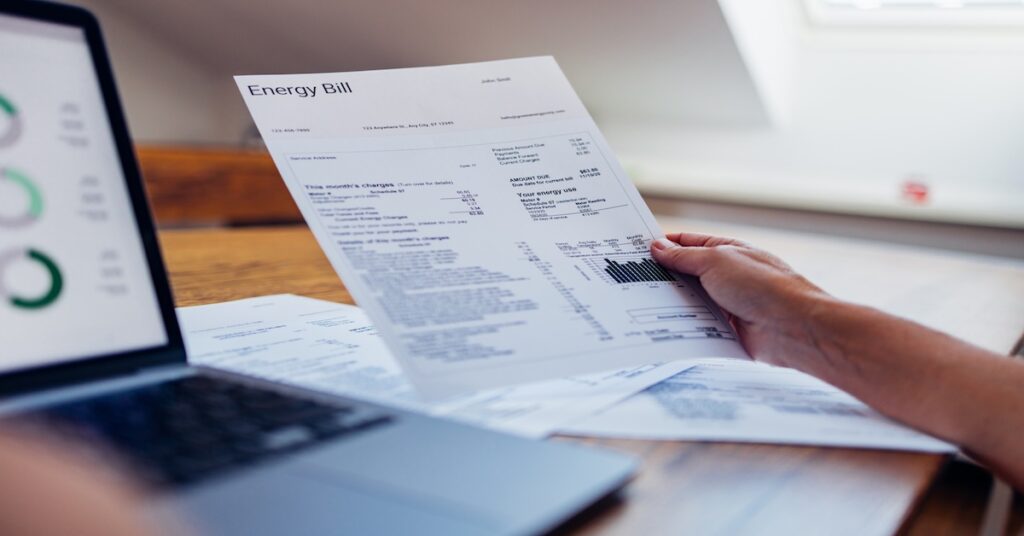 A person reviews their energy bill while using a laptop at home. The document includes various charges and electricity usage information.