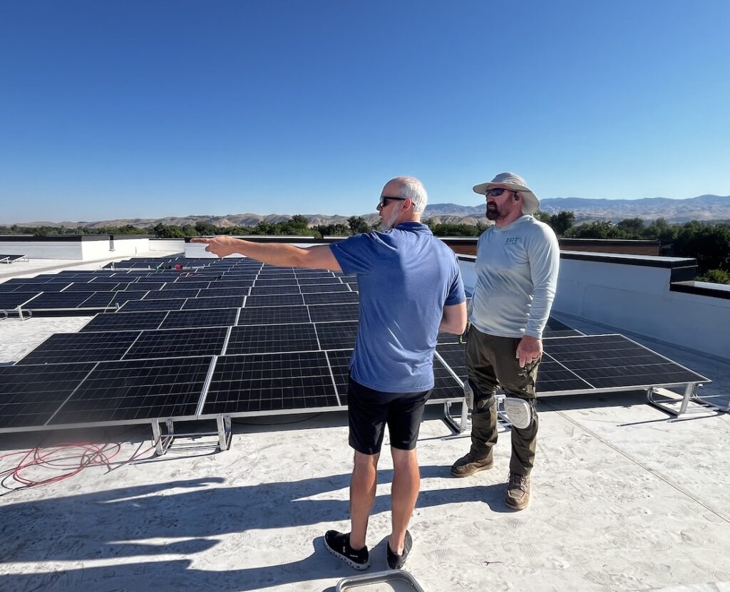 Two solar installers stand on the roof of Whittier Elementary School in Boise, Idaho, with a solar array, a blue sky, and foothills in the background. Both are looking west and one pointing to the other side of the solar array.